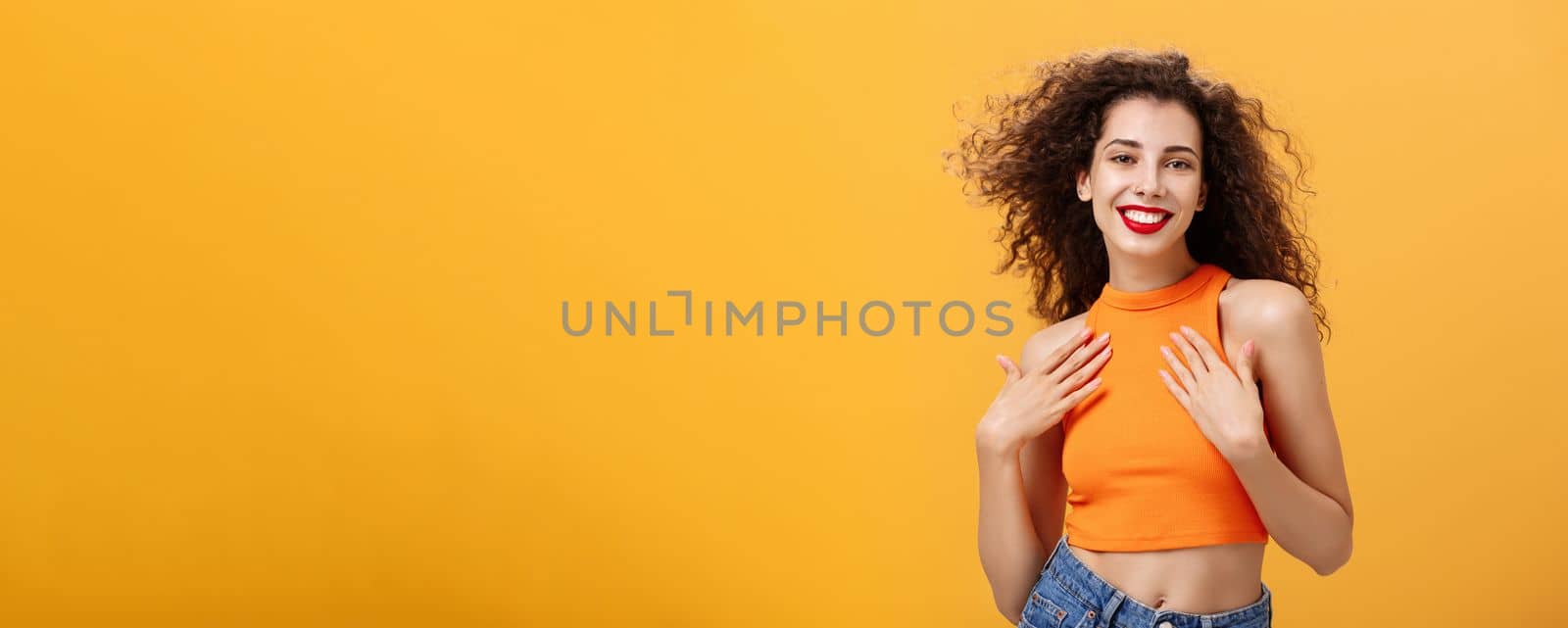 Waist-up shot of sensual and feminine tender female with curly hairstyle in cropped top holding palms on chest delighted and pleased smiling broadly posing flirty and sexy over orange wall by Benzoix