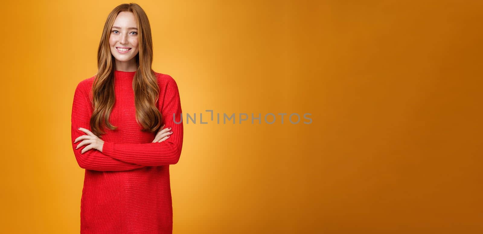 Portrait of elegant and friendly, polite redhead female in red knitted dress holding hands crossed over body in confident and relaxed pose smiling satisfied offering help over orange background.