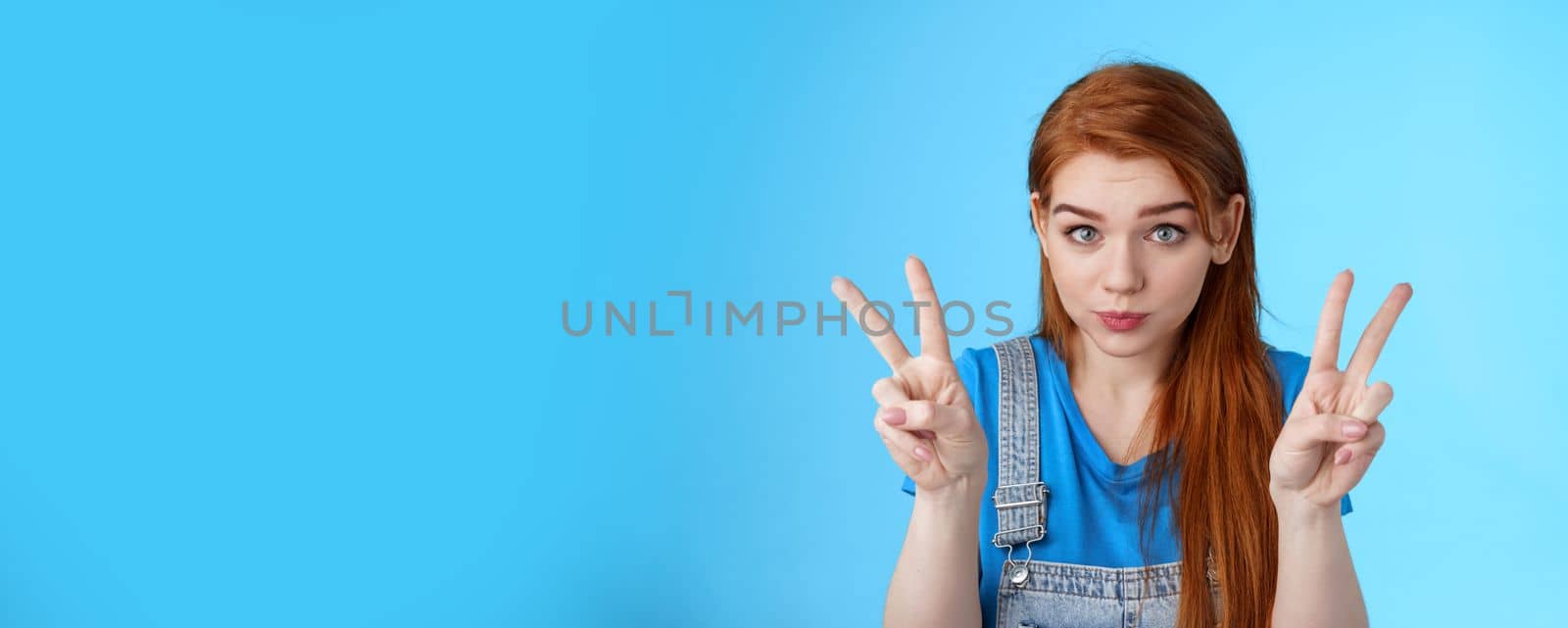 Playful cute silly redhead girlfriend making funny face, show peace victory signs, hold breath, pouting childish, fool around having fun, stand blue background, relaxing entertain siblings.