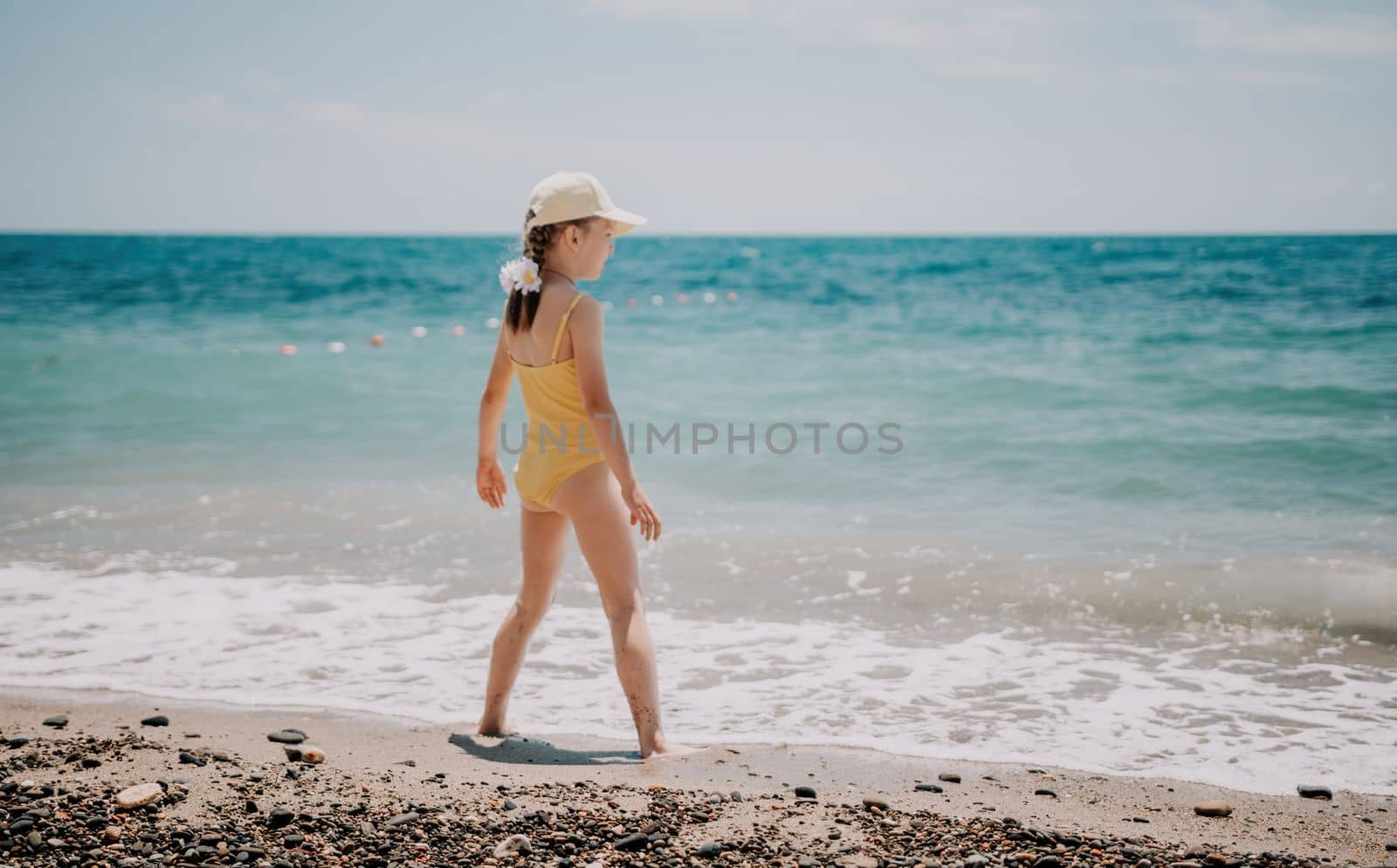 Cute little girl running along the seashore against a clear blue sea and rejoices in the rays of the summer sun. Beautiful girl in yellow swimsuit running and having fun on tropical beach