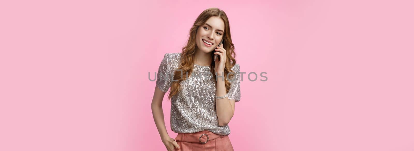 Good-looking sociable european curly-haired woman wearing glitter blouse calling friend discuss party smiling broadly feeling carefree happy, holding smartphone waiting pick up phone by Benzoix