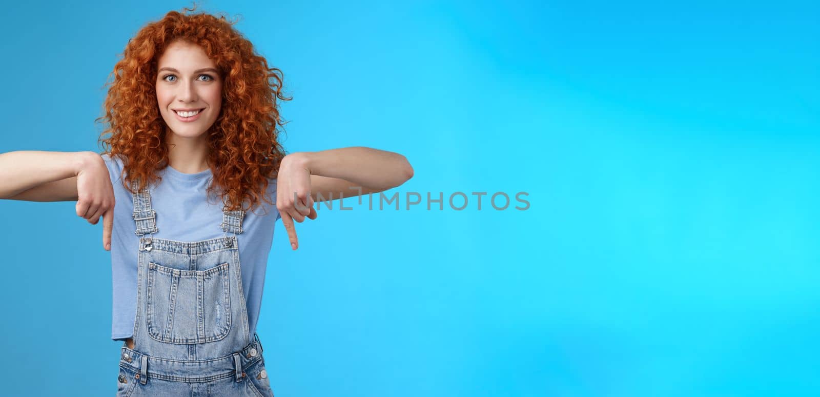 Lifestyle. Charismatic sassy flirty redhead daring ginger girl curly haircut pointing down index fingers smiling broadly enthusiastic explore new store pointing promo like cool advertisement blue background.