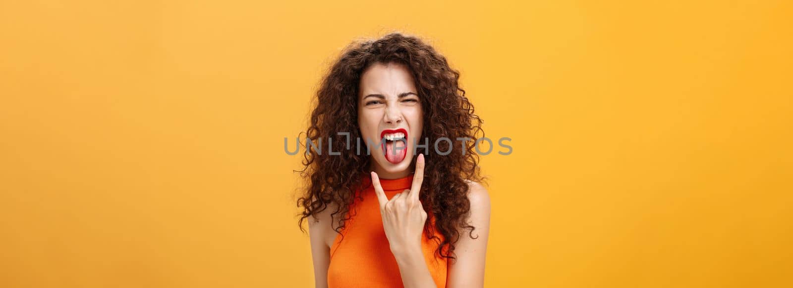 Carefree rebellious stylish urban female with curly hairstyle. sticking out tongue yelling from satisfaction and joy showing rock n roll sign chilling and enjoying cool concert over orange background.