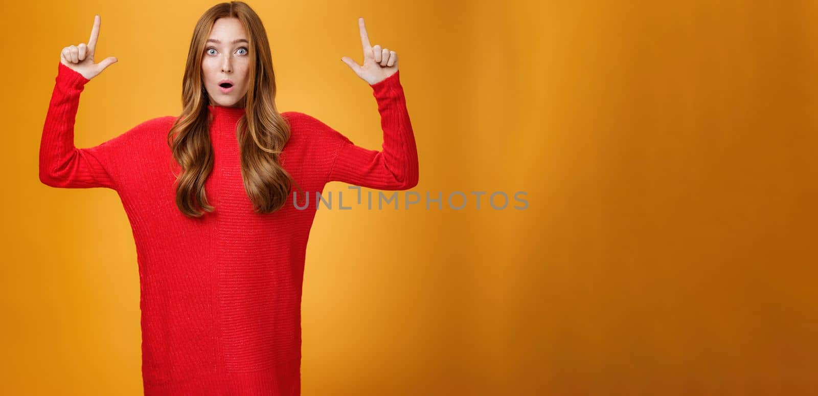 Portrait of attractive and stylish redhead 25s female in knitted red dress gasping from amazement open mouth questioned and surprised as pointing up at astonishing promotion over orange wall by Benzoix