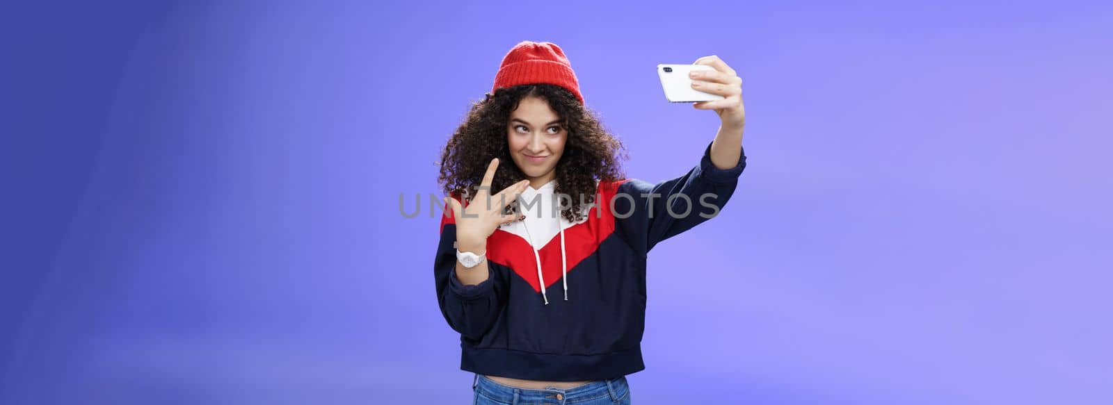 Cool and stylish good-looking caucasian female with curly hair in trendy red beanie and sweatshirt looking from under forehead awesome and swah taking selfie with smartphone holded in extended arm by Benzoix