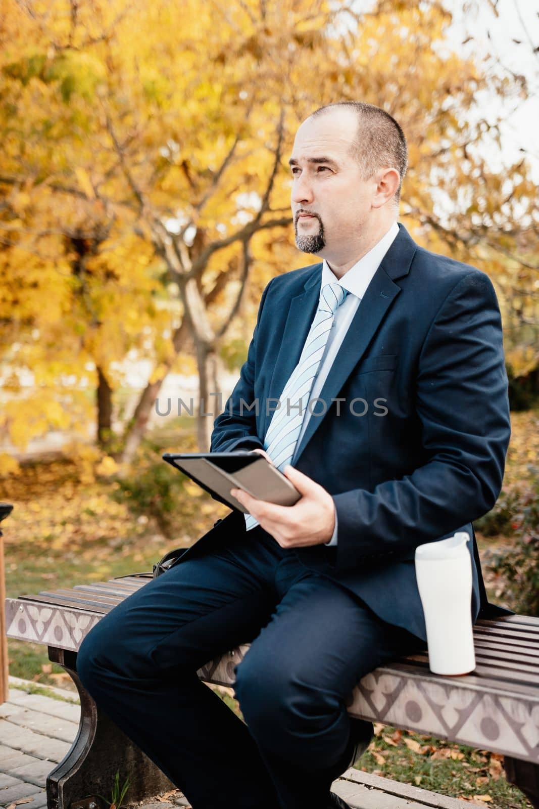 Mature business man in suit reading a news sitting in the park on the bench. Entrepreneur drink coffee in autumn park. Senior executive rest in city park. by panophotograph