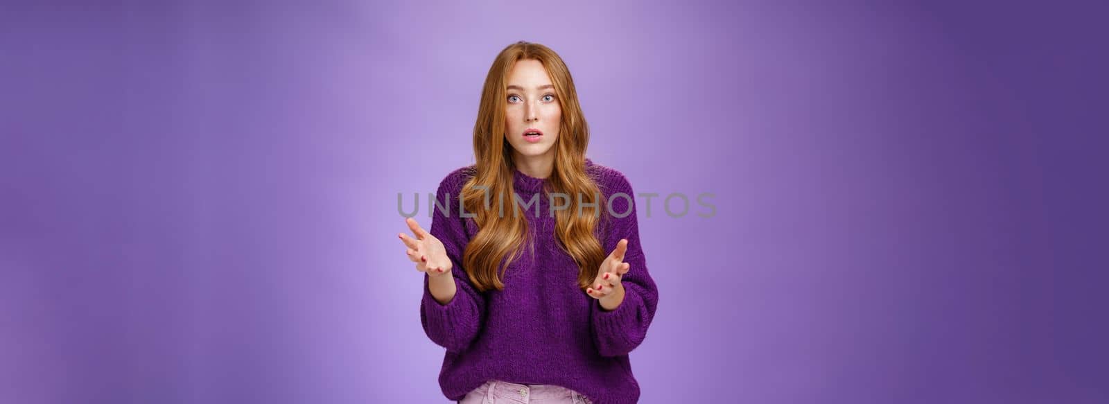 What wrong, I worried. Portrait of nervous and empathical young redhead woman feeling anxious raising hands questioned and looking at camera wondered expressing concern for friend over purple wall.