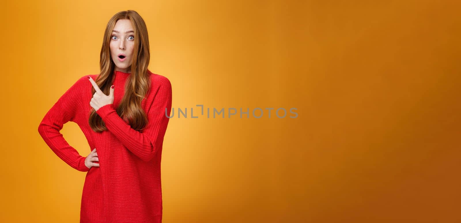Lifestyle. Impressed redhead girl reacting to awesome unbeliavable promotion pointing behind or at upper left corner folding lips staring at camera astonished and surprised over orange background.