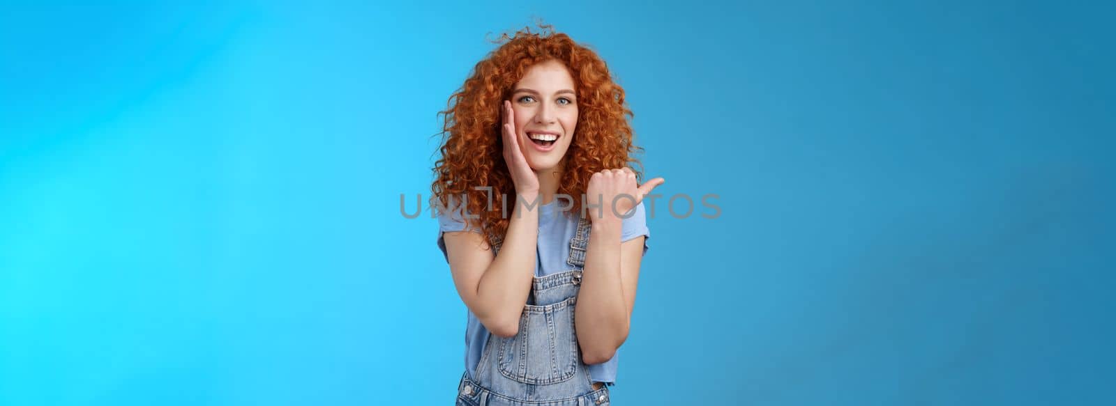 Entertained charismatic good-looking ginger girl curly hairstyle touch cheek gently pleased smiling broadly enthusiastic pointing left thumb impressed excited see awesome promo stunning location by Benzoix