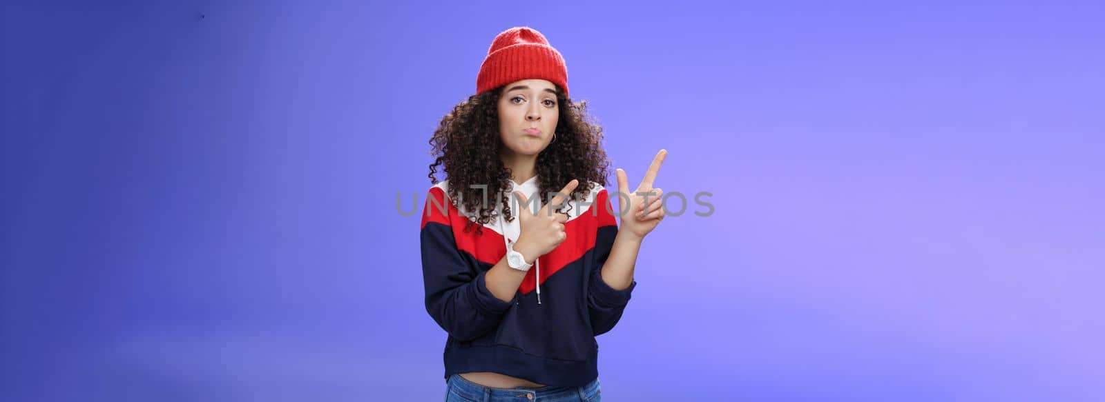 Portrait of silly upset cute curly-haired woman in trendy beanie pursing lips in sadness and frowning making disappointed, regret face pointing at upper left corner unhappy and sad over blue wall by Benzoix