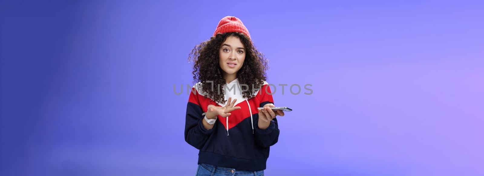 Confused and clueless girl cannot understand meaning of strange message shrugging grimacing questioned as holding smartphone and extending arm in unaware gesture over blue background.