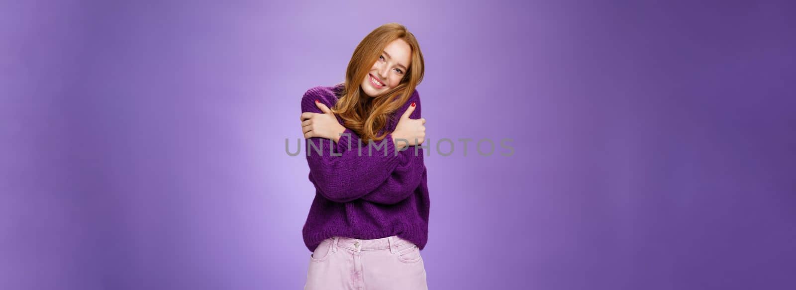 Silly and cute carefree redhead woman in 20s leaning on shoulder as hugging herself feeling warmth wearing purple sweater smiling broadly in cozy and relaxing atmosphere over violet wall by Benzoix