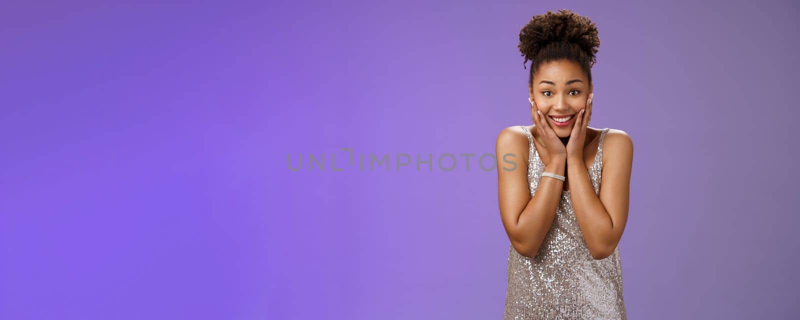 Attractive amused charming fascinated african-american woman in stylish silver dress press hands cheeks astonished grateful receive impressive thrilling gift smiling delighted, blue background.