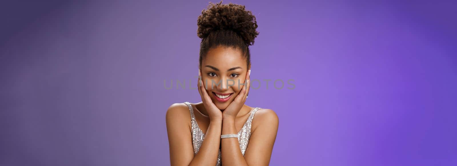 Close-up amused tender romantic african-american woman in stylish glittering silver evening dress touch cheeks blushing cute glad receive passionate compliments standing amused blue background by Benzoix