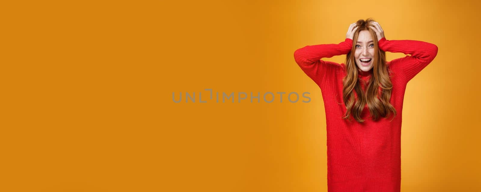 Excited and overwhelmed charming redhead woman in knitted warm red dress yelling from thrill and happiness holding hands on hair smiling broadly impressed with unexpected luck over orange wall by Benzoix