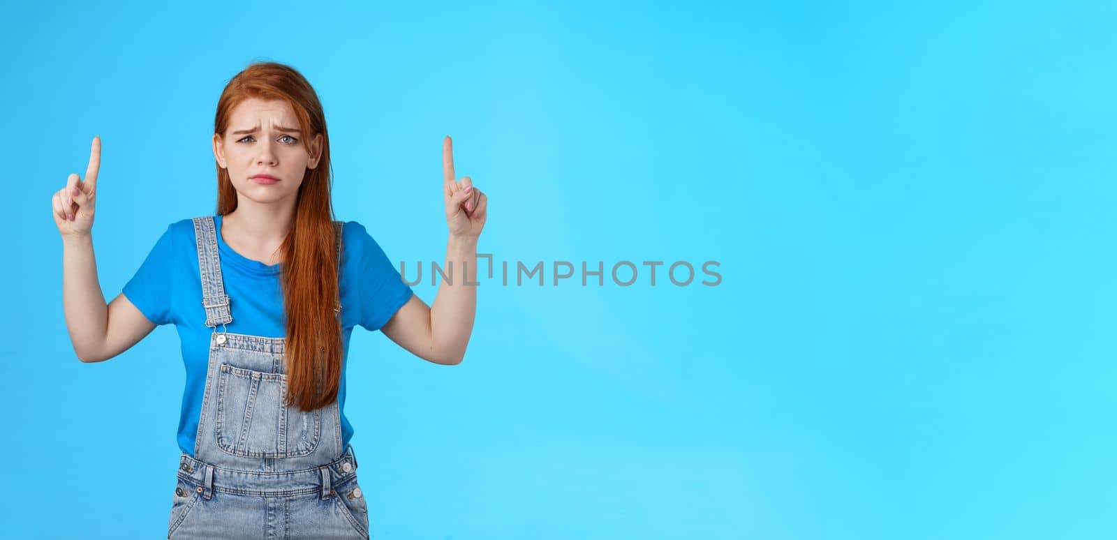 Doubtful concerned unsure redhead female making important decision, grimacing hesitant displeased, point up promo, top copy space, feel uncertain unlikely buy suspicious product, blue background.