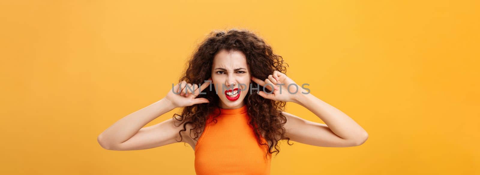 Waist-up shot of irritated bothered stylish woman. with curly hair and red lipstick squinting clenching teeth waiting for loud bang covering ears with index fingers being annoyed of noisy neighbours.