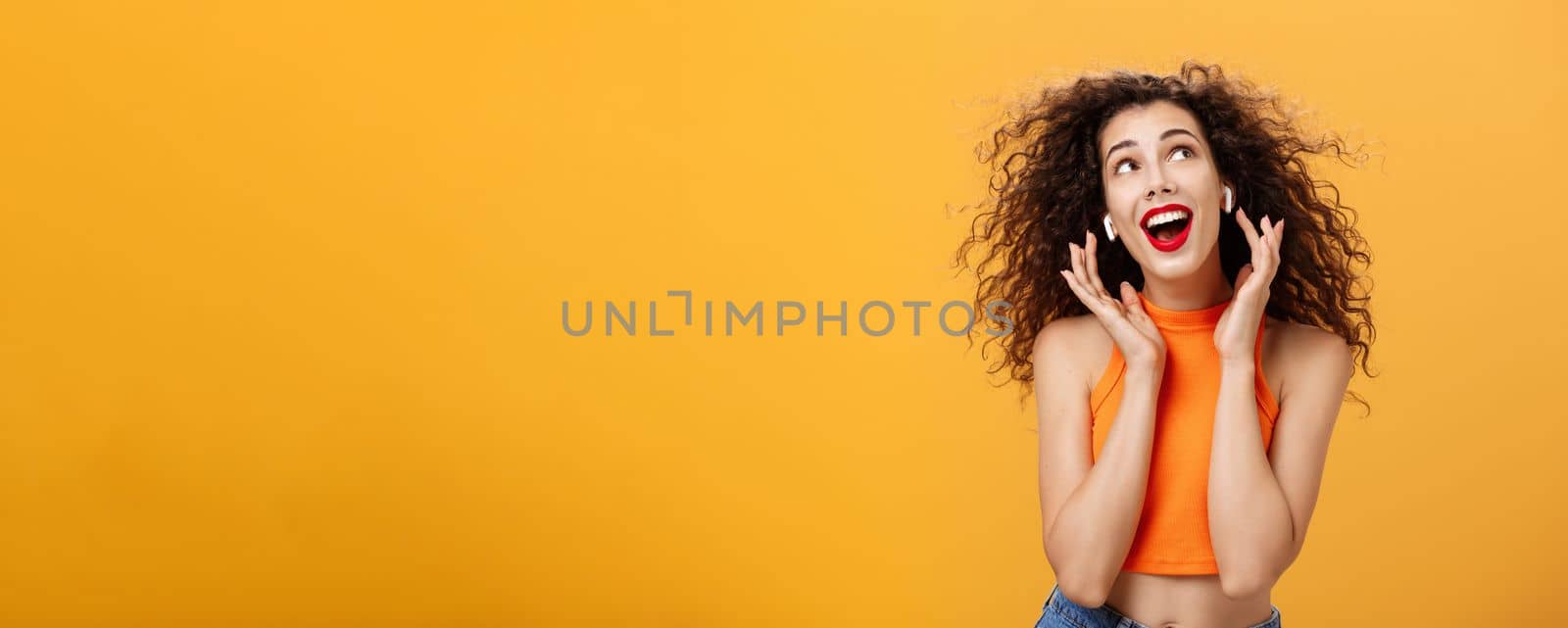 Waist-up shot of attractive silly curly-haired european female. in cropped top using wireless earphones touching earbuds and gazing at upper right corner delighted and carefree over orange wall.