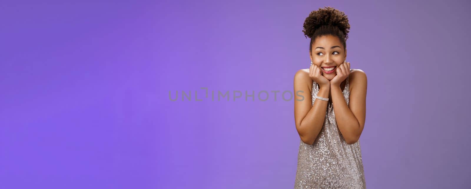 Amused silly cute black girl in silver dress. anticipating exciting party dreamy gaze up touch cheeks smiling broadly optimistic waiting dream come true standing pleased touched blue background.