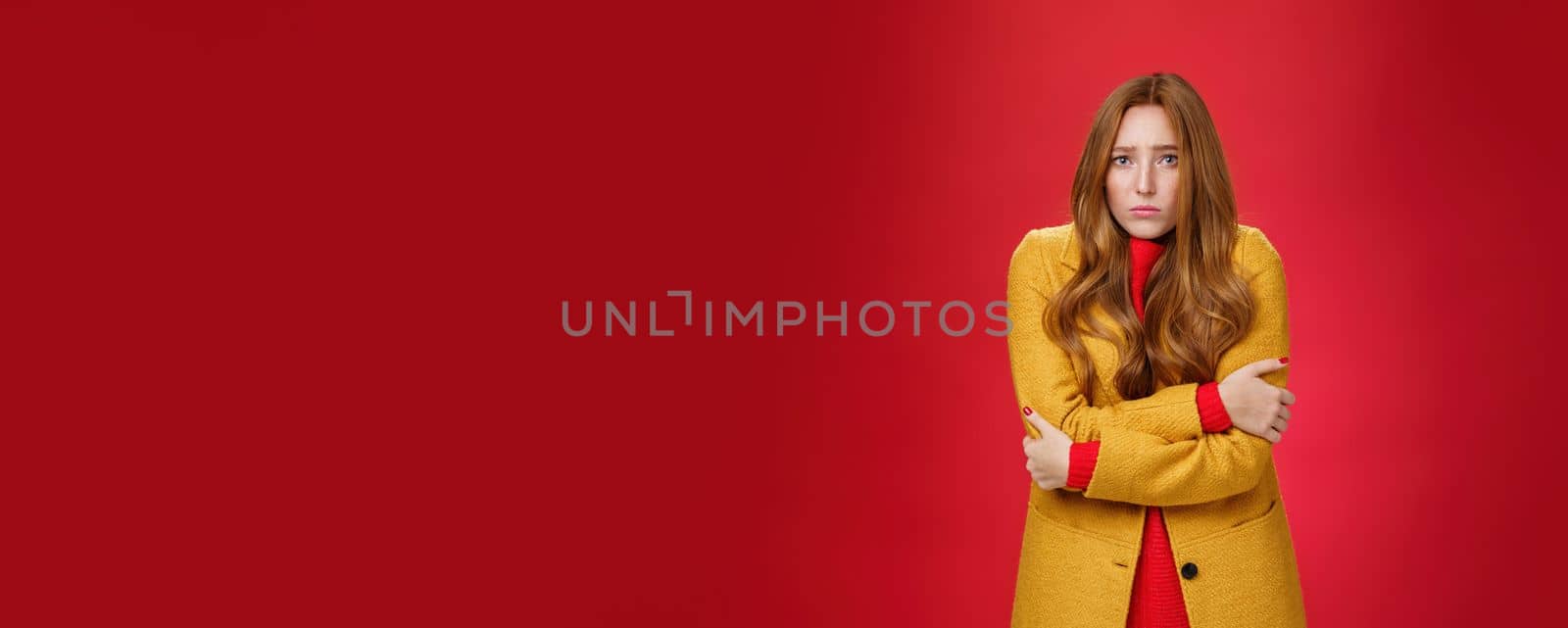 Cute and sad trembling redhead girl in light yellow coat shaking from cold, having goosebumps frowning and pouting from sorrow as freezing on windy weather over red background. Copy space