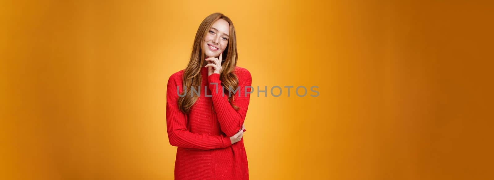 Sweet and tender redhead woman with cute freckles in red knitted dress tilting head touching face with fingers and smiling delighted and loving a camera over orange background. Copy space