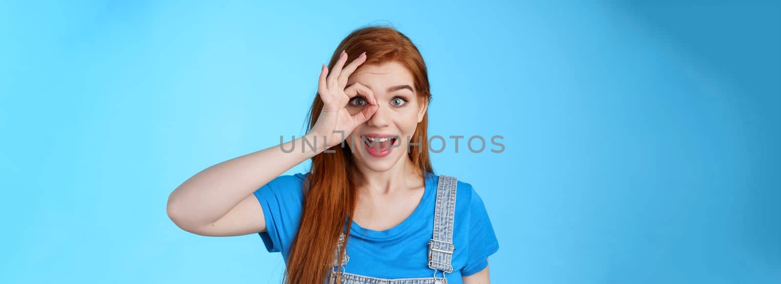 Dreamy curious attractive cheerful redhead woman surprised, look fascinated camera look through fingers okay sign, stand amused, gaze admiration blue background, check out promo by Benzoix