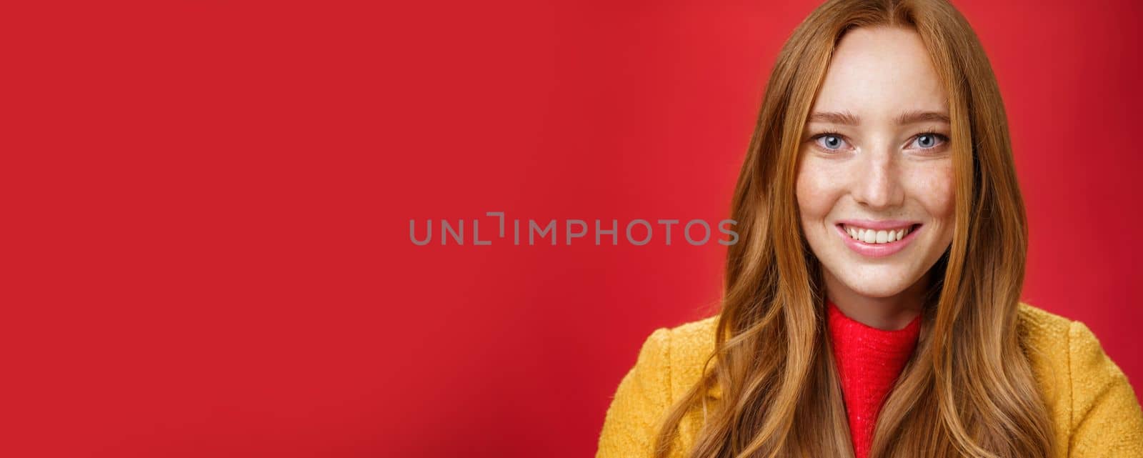 Headshot of hopeful and happy young redhead 20s girlfriend with cute freckles and blue eyes smiling broadly with faithful excited expression having high hopes, posing over red background. Emotions concept