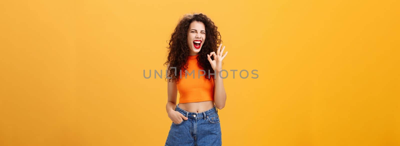 Excellent party like it. Joyful enthusiastic young curly-haired female with red lipstick in stylish cropped top winking excited smiling and showing okay or perfect sign posing over orange background.