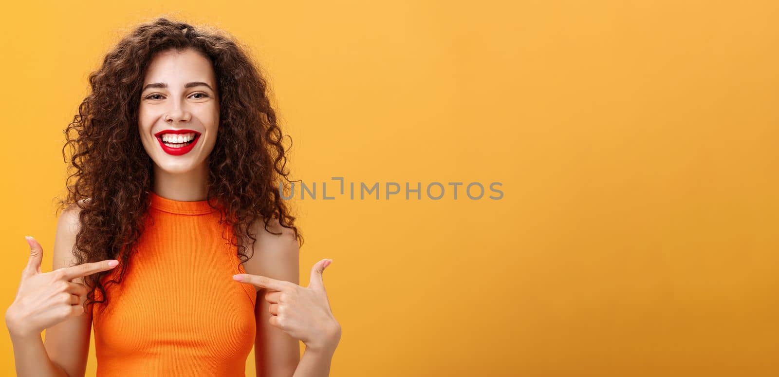 Waist-up shot of proud and happy outgoing charming woman with curly hair and red lipstick pointing at herself delighted and confident talking about own achievement over orange background. Copy space