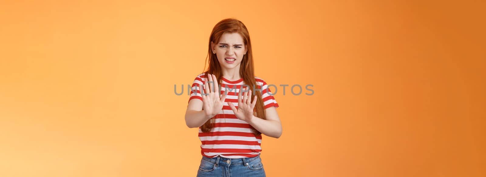 Gosh it stinks. Disgusted redhead picky woman blocking sign raise hands up defensive, grimacing, cringe from aversion awful smell, show refusal rejecting disgusting offer, stand orange background by Benzoix