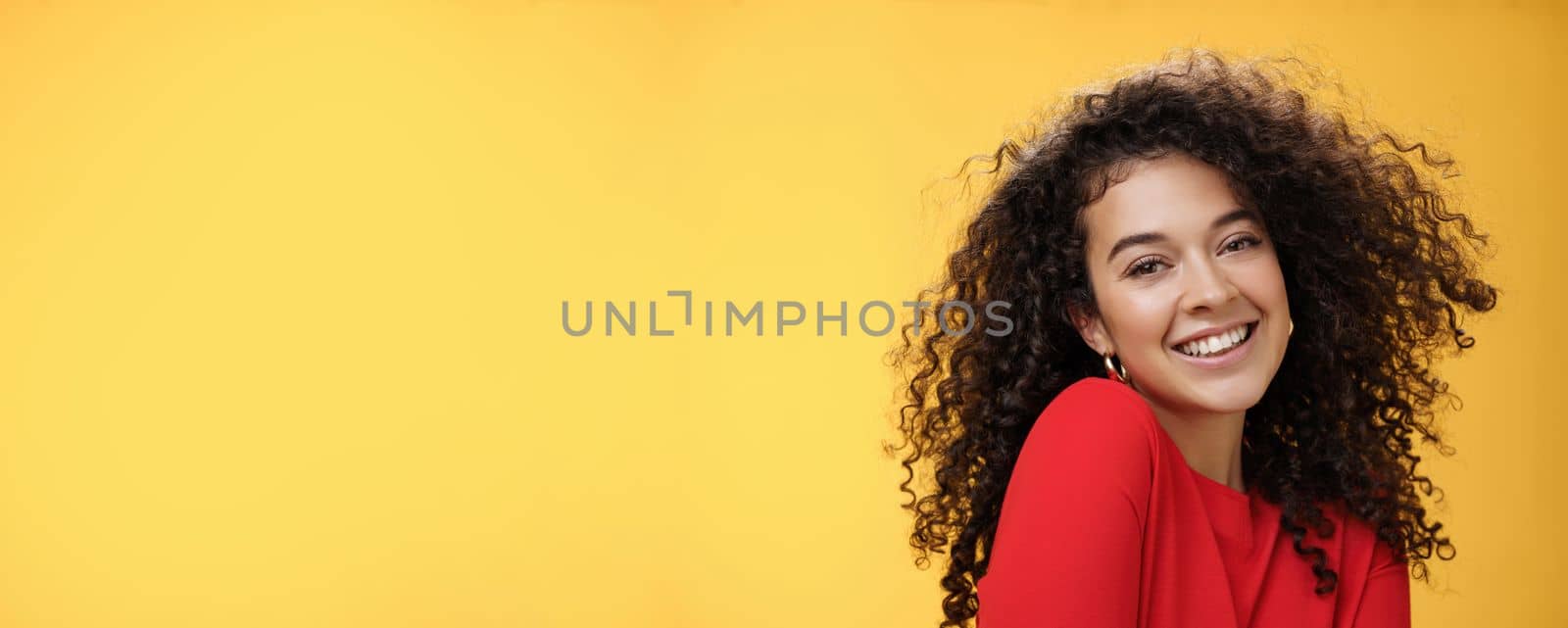 Romantic and flirty young cute caucasian girlfriend with curly hair in red blouse making silly pose smiling joyfully and lifting shoulder coquettish smiling broadly at camera over yellow wall by Benzoix