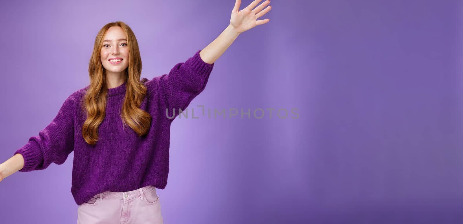 Girl stretched hands sideways to welcome and greet friend giving warm hug smiling broadly at camera standing joyful wanting cuddle over purple background, wearing violet sweater and pants by Benzoix