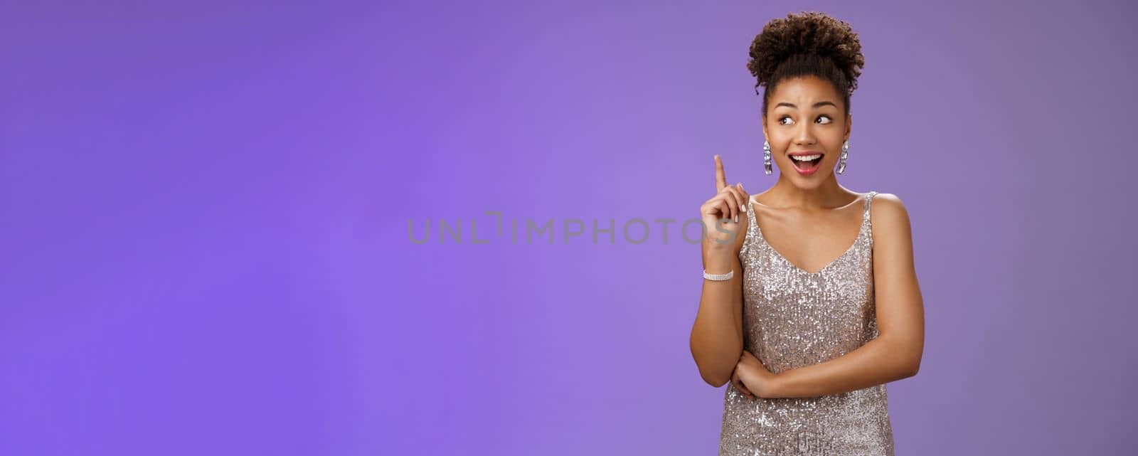 Creative joyful dreamy young african-american girl in silver dress raise index finger eureka gesture look aside inspired have awesome idea sharing suggestion tell plan aloud, standing blue background.