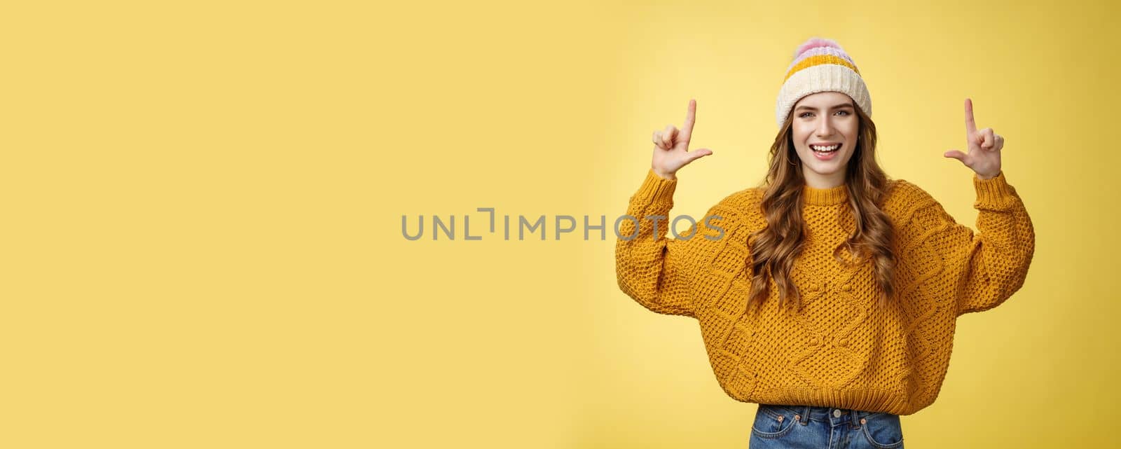 Friendly charming energized young 20s woman dress-up stylish hat sweater having fun introducing promotion pointing raised index finger up showing top advertisement, smiling happily yellow background.