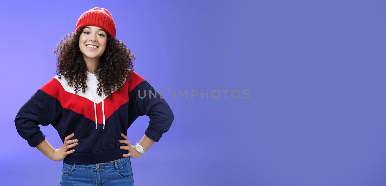 Confident ad energized cute european woman with curly hairstyle in warm beanie and stylish sweatshirt holding hands on waist and smiling friendly at camera as ready to go out and have fun outside by Benzoix