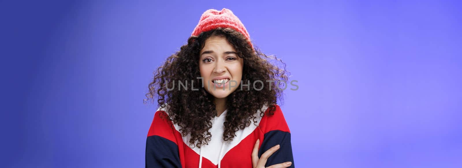 Woman with curly hair in winter beanie feeling uncomfortable and discomfort clenching teeth and frowning intense as hugging herself insecure and awkward, unwilling to say cruel rejection, feel awkward by Benzoix