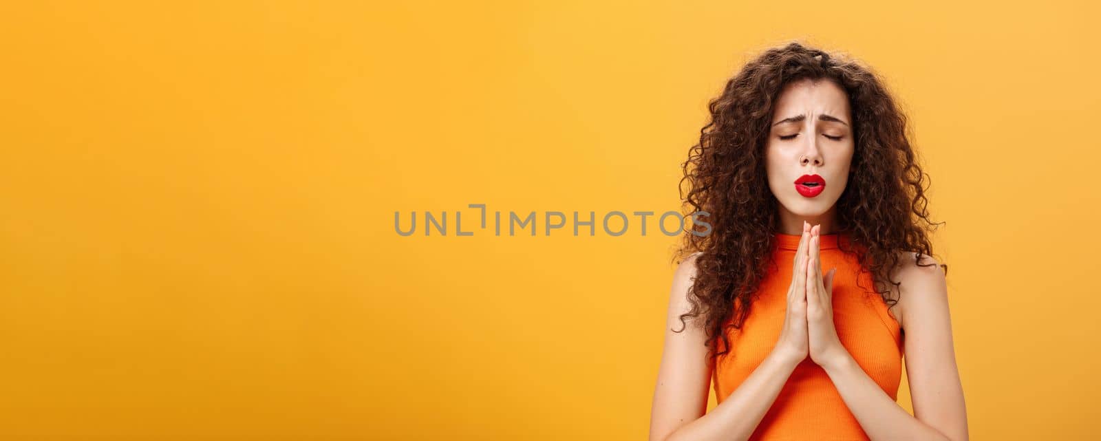 Nervous and concerned woman. with curly hairstyle feeling hopeful praying with closed eyes and frowned eyebrows holding hands in pray near chest hopefully dreaming troubled will solve over orange wall. Emotions and body language concept