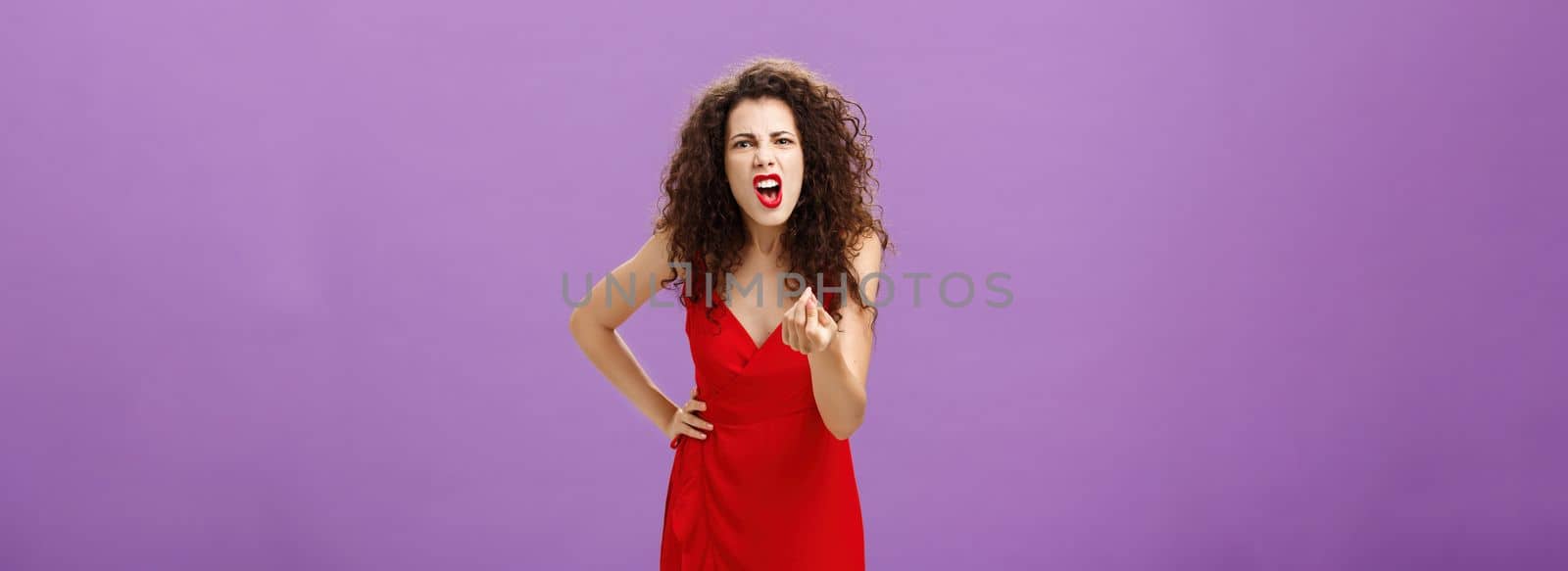 Woman in fury fighting with husband over money. making italian gesture with fingers shouting and yelling from fury and anger standing displeased and pissed over purple wall in red elegant dress.