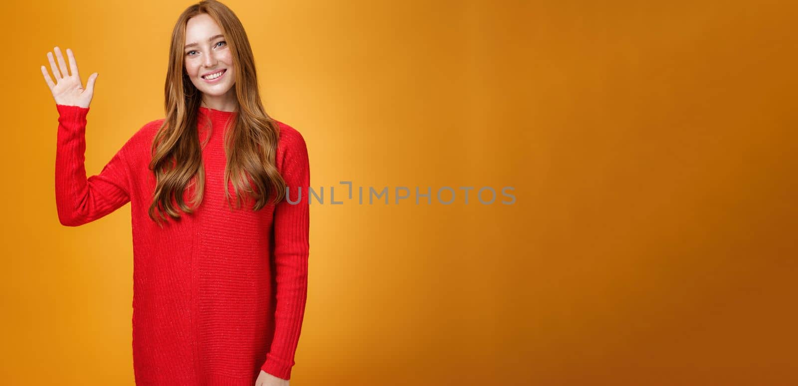 Friendly and optimistic good-looking ginger girl in red sweater raising palm waving at camera, saying hello or hi joyfully smiling cute, greeting new members posing over orange background.