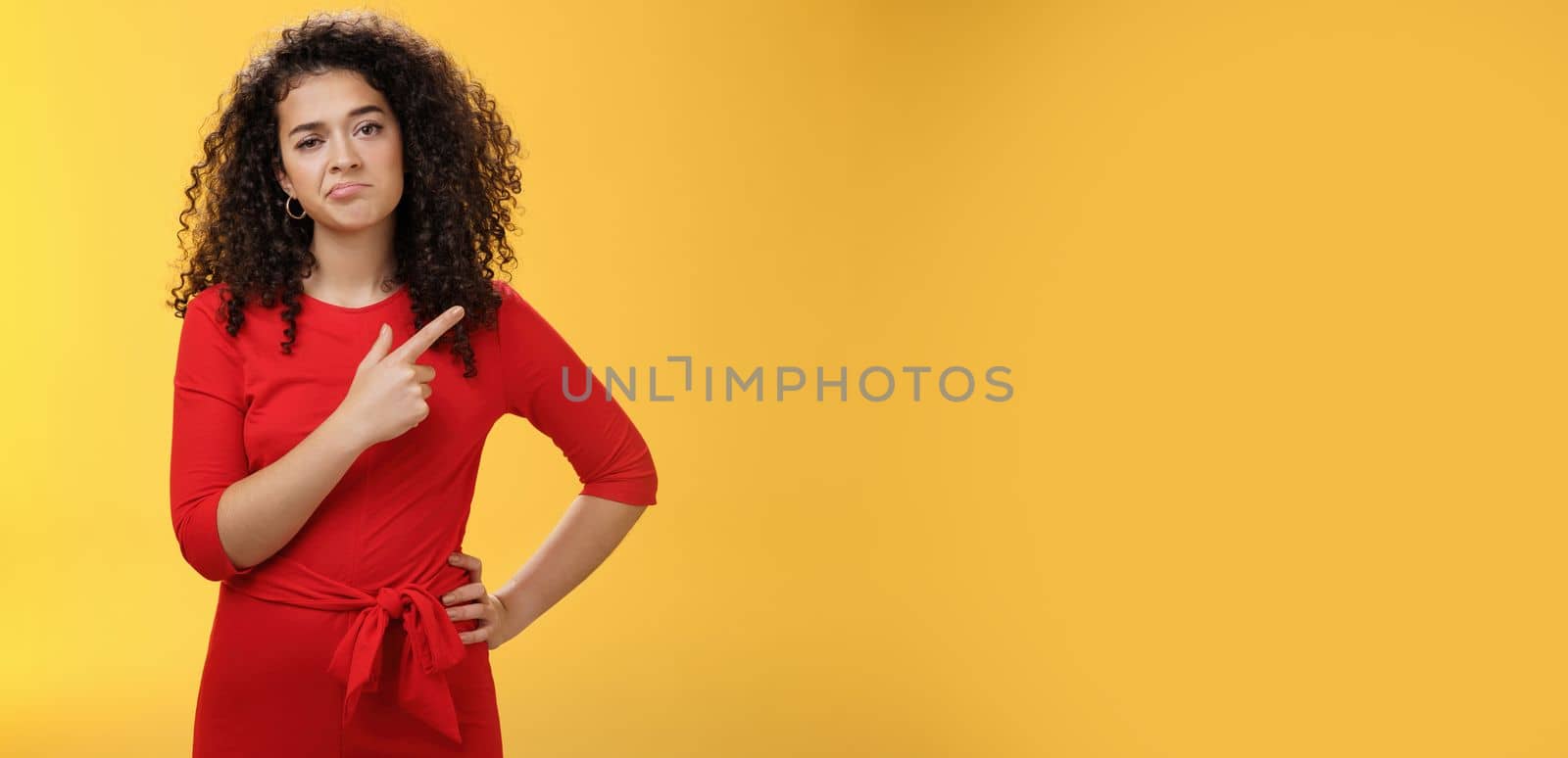 Portrait of unimpressed careless and bored young curly-haired female coworker in dress making upset smile pointing at upper right corner looking with indifference and disappointment. Body language concept