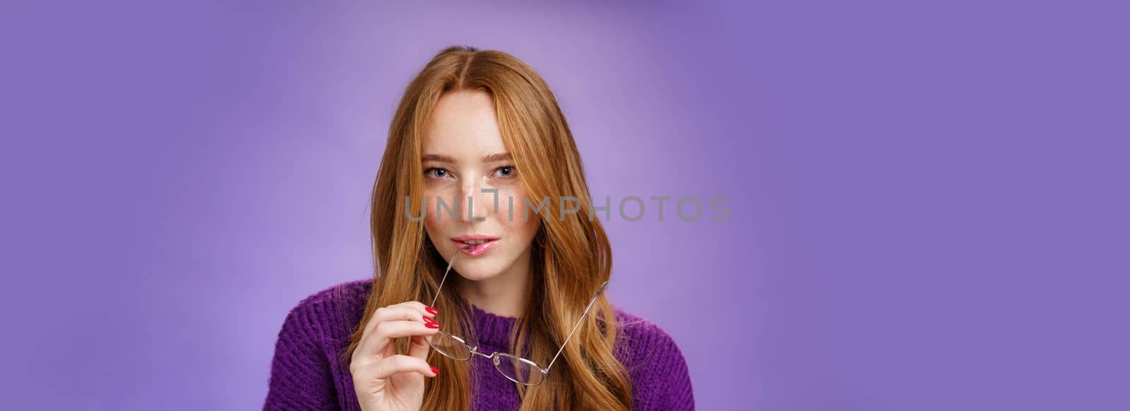 Close-up shot of smart and creative female redhead genius biting frame of glasses and squinting curious at camera as thinking having idea, making up plan against purple background.