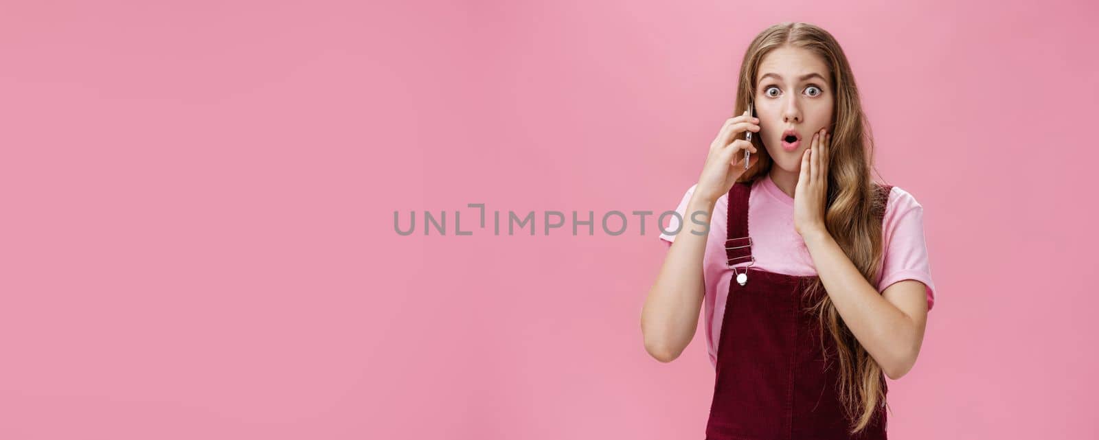 Waist-up shot of shocked woman learning stunning news via smartphone talking on phone holding cellphone near ears folding lips and gasping in surprise popping eyes at camera worried over pink wall by Benzoix