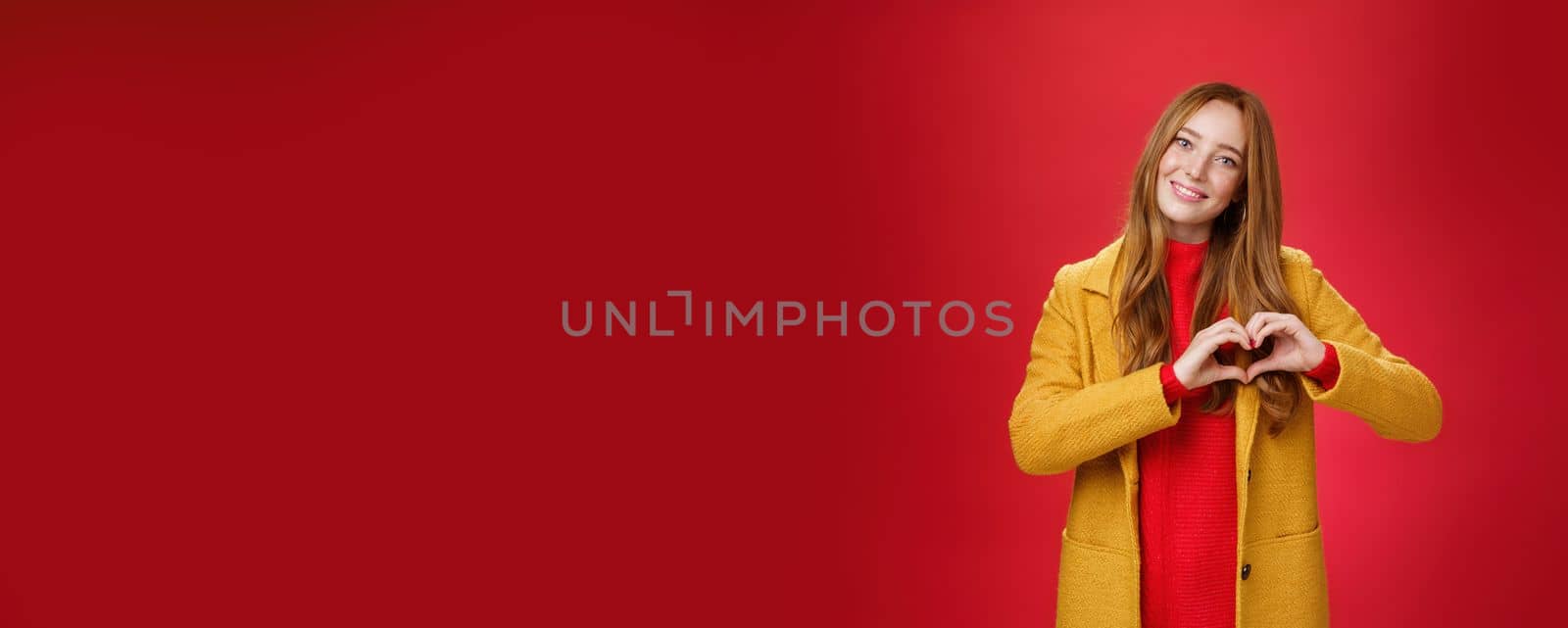 Love, romance and fall concept. Portrait of charming tender and gentle young redhead woman in yellow coat showing heart gesture making confession in sympathy, smiling cute at camera over red wall.