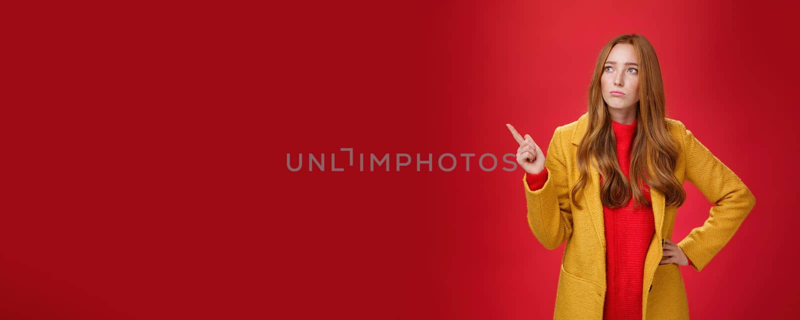 Sad and gloomy cute ginger girl in 20s wearing trendy yellow coat holding hand on waist, pouting, looking and pointing at upper left corner with regret and sorrow over red background by Benzoix