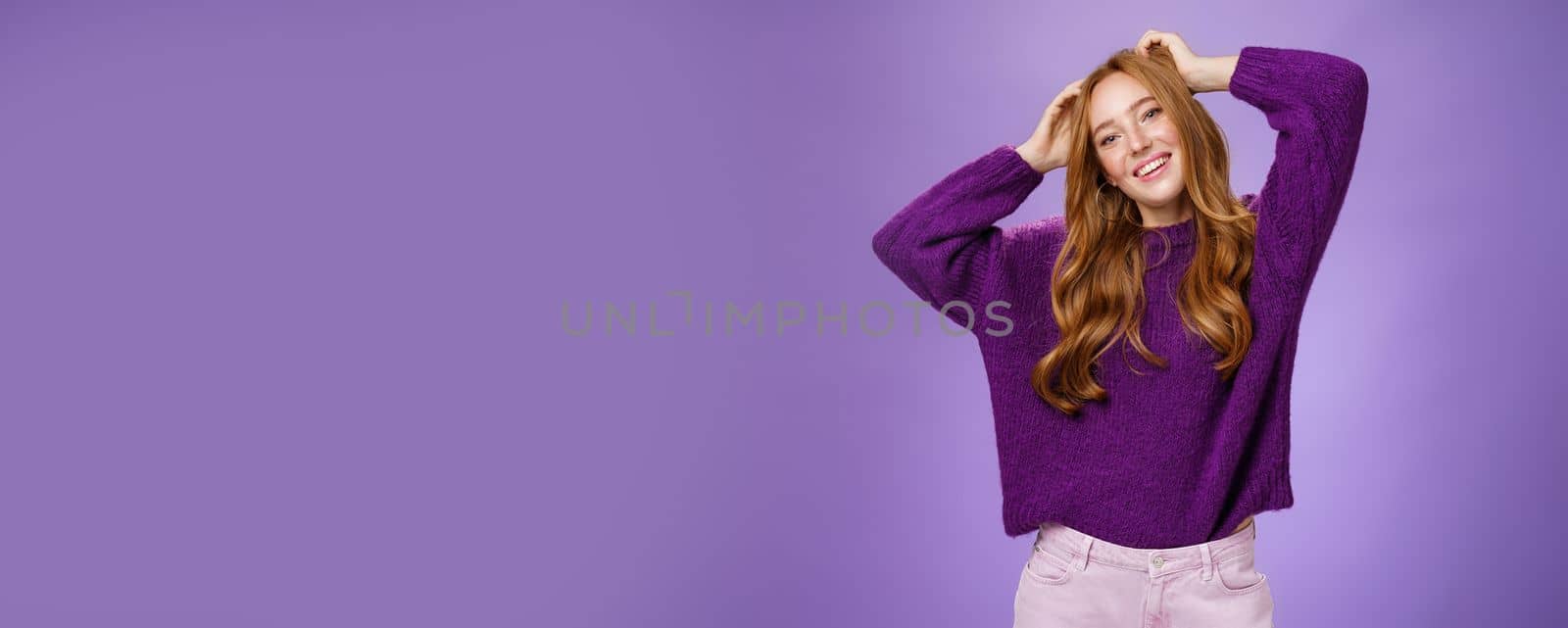 Carefree joyful and cool stylish redhead female student in purple warm sweater enjoying holidays stretching, holding hands near head, tilting and smiling happily relaxing against violet background.