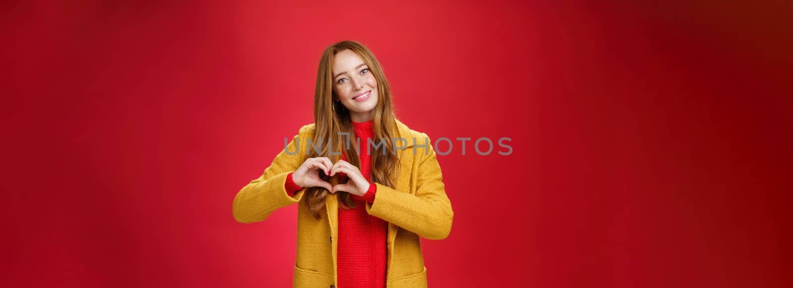Love, romance and fall concept. Portrait of charming tender and gentle young redhead woman in yellow coat showing heart gesture making confession in sympathy, smiling cute at camera over red wall.