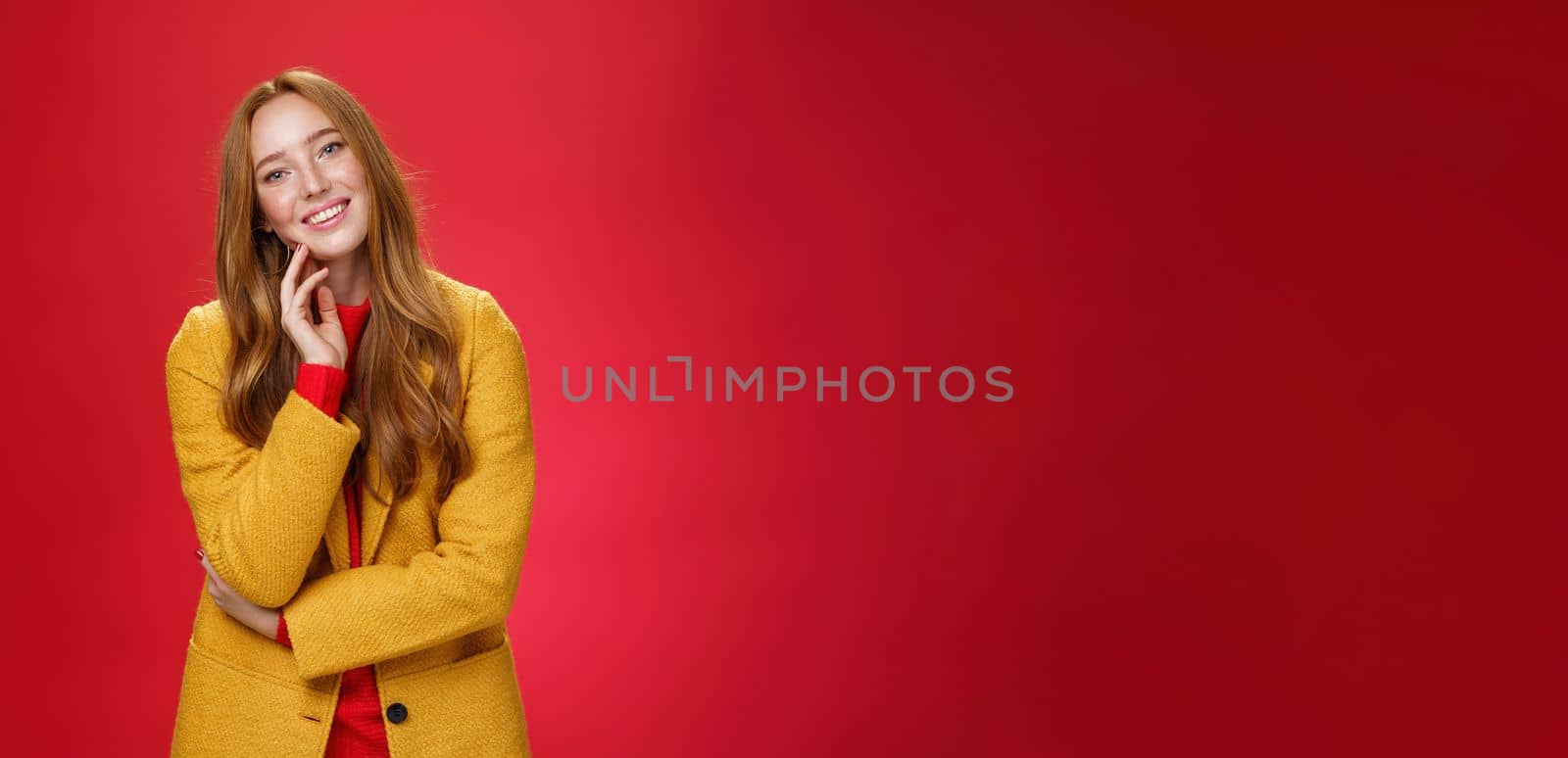 Good-looking carefree and happy relaxed redhead girlfriend in yellow stylish coat touching face and tilting head as smiling with positive emotions, posing cheerful against red background.