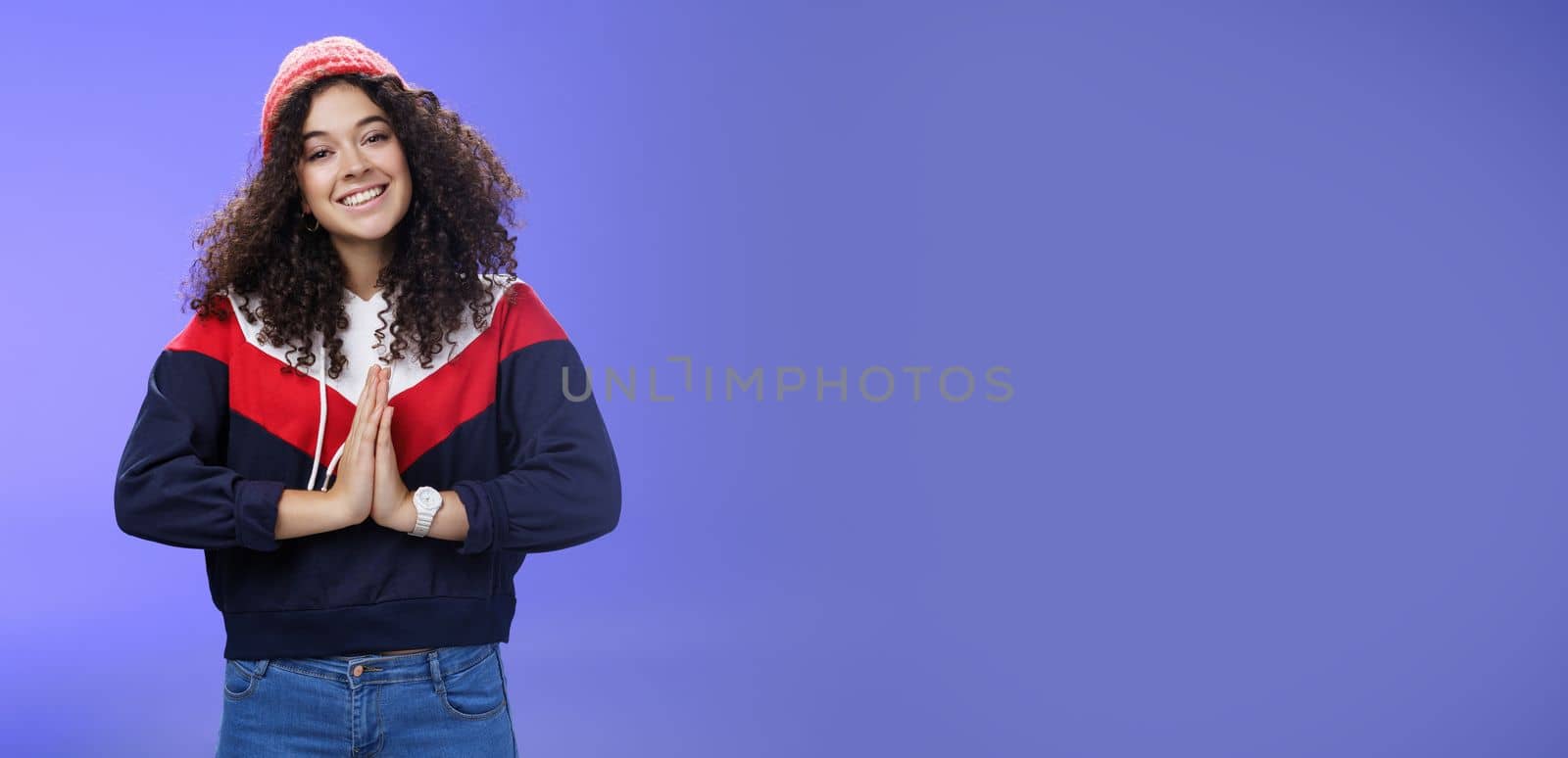 Portrait of charming curly-haired feminine girlfriend begging for favour as posing over blue background in winter outfit with hat, holding hands in pray smiling with angel look and friendly gaze. Body language and facial expressions concept