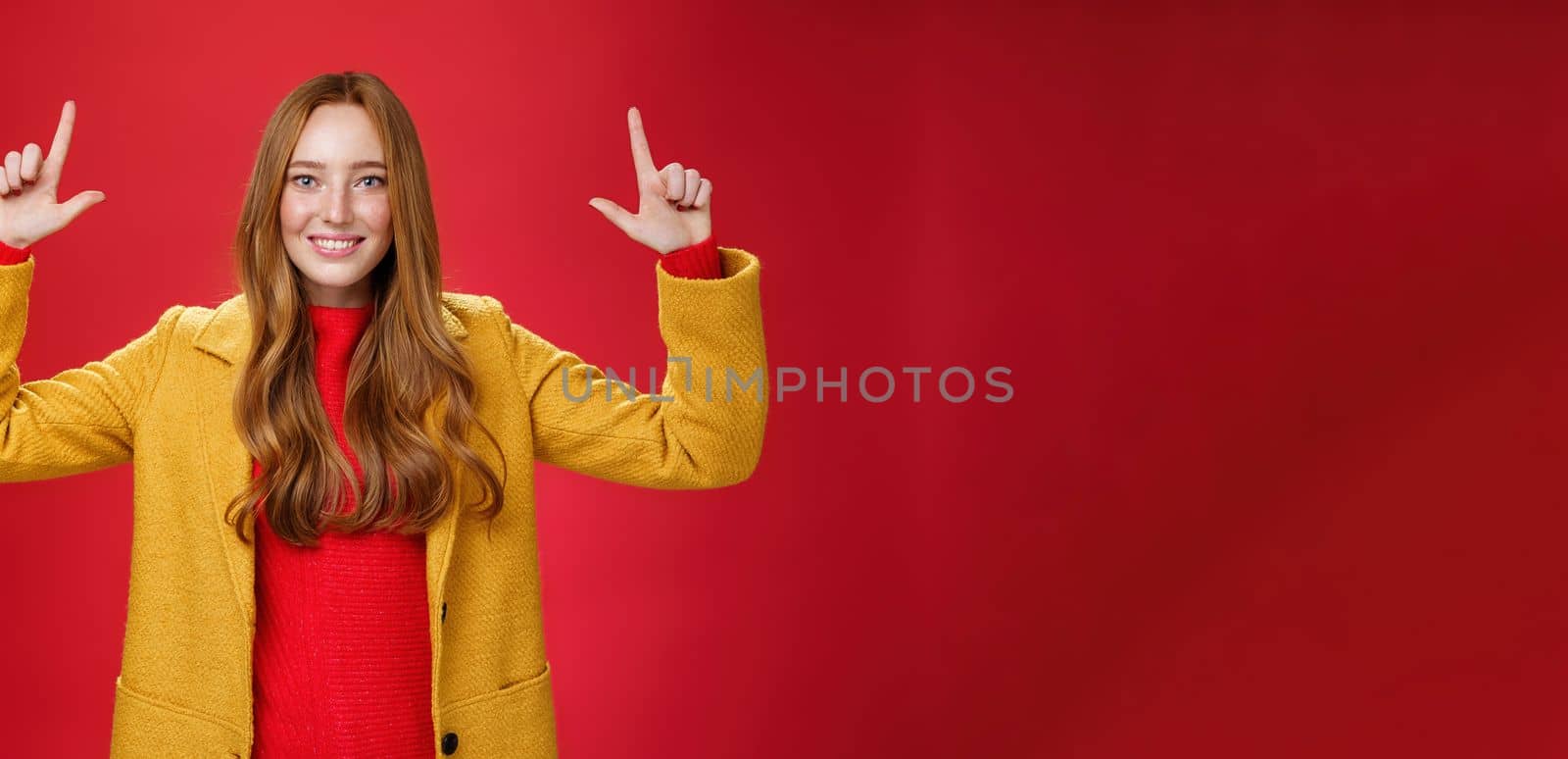 Indoor shot of redhead attractive woman in yellow fall coat raising hands promoting advertisement as pointing up and smiling broadly with satisfied pleasant expression over red background by Benzoix