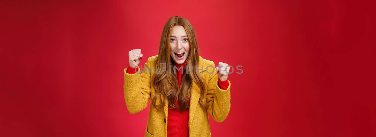Studio shto of cheerful excited good-looking ginger girl in yellow coat raising clenched fists in joy and happiness, triumphing yelling yes from success and triumph, celebrating win over red wall.